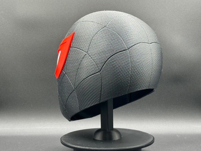 Puede incluir: Una m&aacute;scara impresa en 3D de color negro y rojo con un dise&ntilde;o de telara&ntilde;a. La m&aacute;scara est&aacute; en un soporte negro.