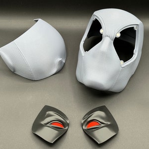 Może przedstawiać: Zestaw pięciu element&oacute;w plastikowej maski w kolorze białym i czarnym. Maska składa się z pięciu element&oacute;w: gł&oacute;wnego elementu maski, dw&oacute;ch element&oacute;w oczu i dw&oacute;ch element&oacute;w bocznych. Elementy oczu mają czerwone oczy.