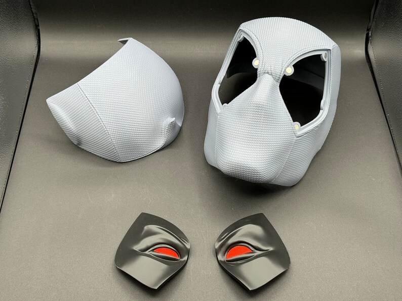 Może przedstawiać: Zestaw pięciu element&oacute;w plastikowej maski w kolorze białym i czarnym. Maska składa się z pięciu element&oacute;w: gł&oacute;wnego elementu maski, dw&oacute;ch element&oacute;w oczu i dw&oacute;ch element&oacute;w bocznych. Elementy oczu mają czerwone oczy.