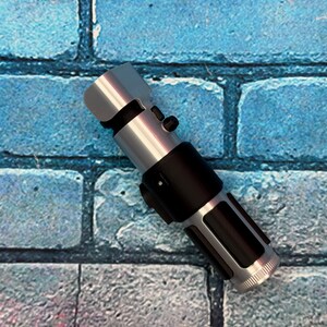 May include: A silver and black lightsaber prop, featuring a cylindrical design with detailed metallic accents. The hilt is black, and the blade emitter is silver. The lightsaber is set against a blue brick wall background.