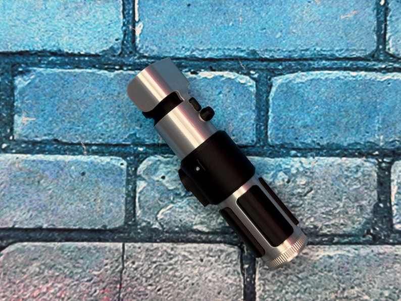 May include: A silver and black lightsaber prop, featuring a cylindrical design with detailed metallic accents. The hilt is black, and the blade emitter is silver. The lightsaber is set against a blue brick wall background.