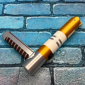 May include: A gold and silver lightsaber prop with white and silver accents. The hilt is silver with a black button. The blade is gold. The prop is lying on a blue brick surface.