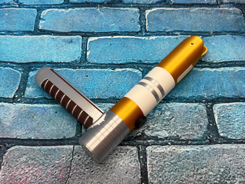 May include: A gold and silver lightsaber prop with white and silver accents. The hilt is silver with a black button. The blade is gold. The prop is lying on a blue brick surface.
