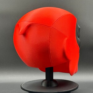 Puede incluir: M&aacute;scara de Deadpool de pl&aacute;stico rojo con una superficie texturizada. La m&aacute;scara est&aacute; en un soporte negro.