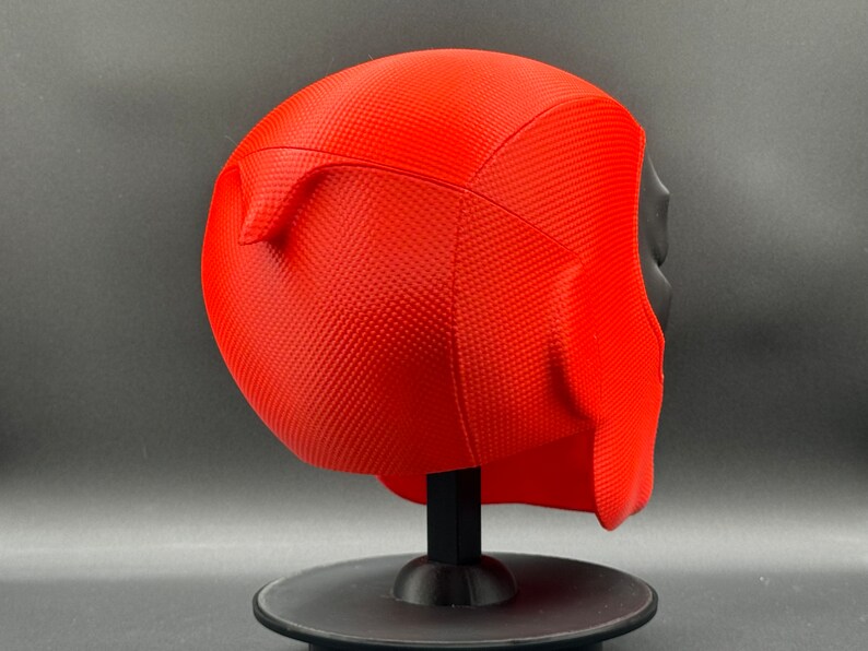 Puede incluir: M&aacute;scara de Deadpool de pl&aacute;stico rojo con una superficie texturizada. La m&aacute;scara est&aacute; en un soporte negro.