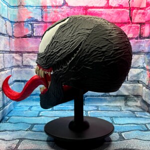 May include: A black Venom mask with a textured surface, white eyes, and a long red tongue. The mask is displayed on a black stand with a round base. The mask has sharp teeth and is a replica of the Marvel Comics character.