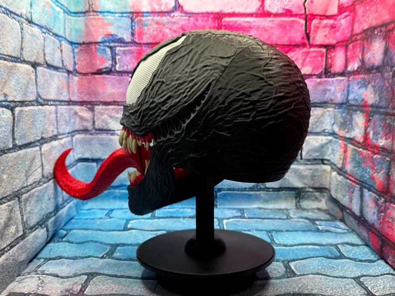 May include: A black Venom mask with a textured surface, white eyes, and a long red tongue. The mask is displayed on a black stand with a round base. The mask has sharp teeth and is a replica of the Marvel Comics character.