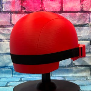 May include: A smooth, bright red, rounded helmet-like headpiece featuring a horizontal black elastic strap that wraps around the circumference. The object is mounted on a black circular stand, showcasing a clean, solid-colored, minimalist design.