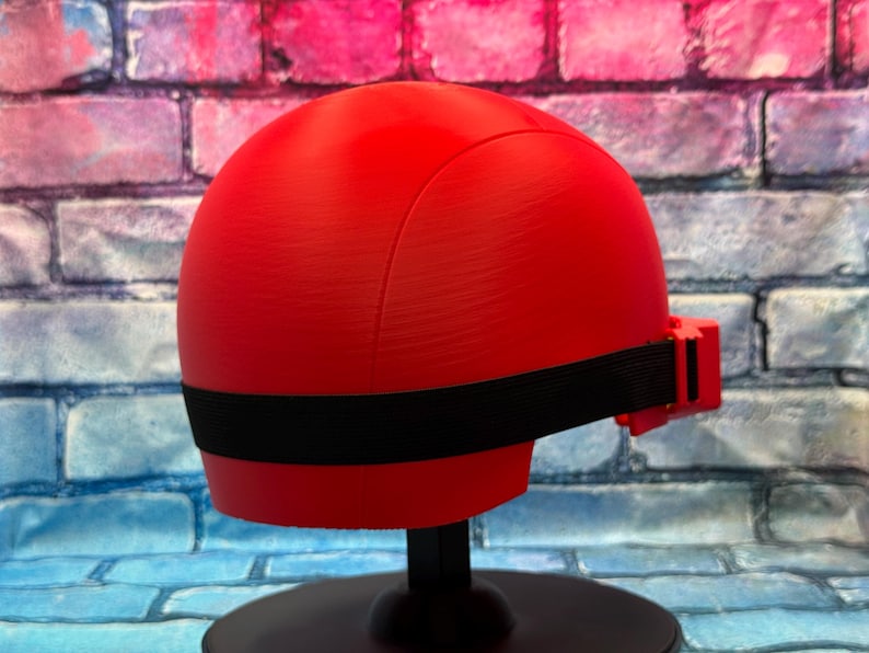 May include: A smooth, bright red, rounded helmet-like headpiece featuring a horizontal black elastic strap that wraps around the circumference. The object is mounted on a black circular stand, showcasing a clean, solid-colored, minimalist design.
