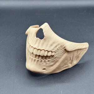Puede incluir: Una m&aacute;scara de calavera beige que cubre la mitad inferior de la cara, con dientes detallados y una cavidad nasal visible. La m&aacute;scara tiene una superficie texturizada y probablemente se utiliza para disfraces o decoraci&oacute;n.