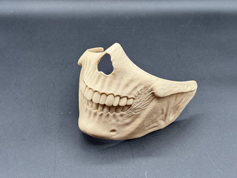 Puede incluir: Una m&aacute;scara de calavera beige que cubre la mitad inferior de la cara, con dientes detallados y una cavidad nasal visible. La m&aacute;scara tiene una superficie texturizada y probablemente se utiliza para disfraces o decoraci&oacute;n.