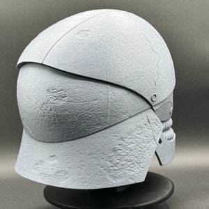 May include: A white, textured helmet with a segmented design. The helmet has a rounded top and a pointed chin guard. The helmet is on a black stand.