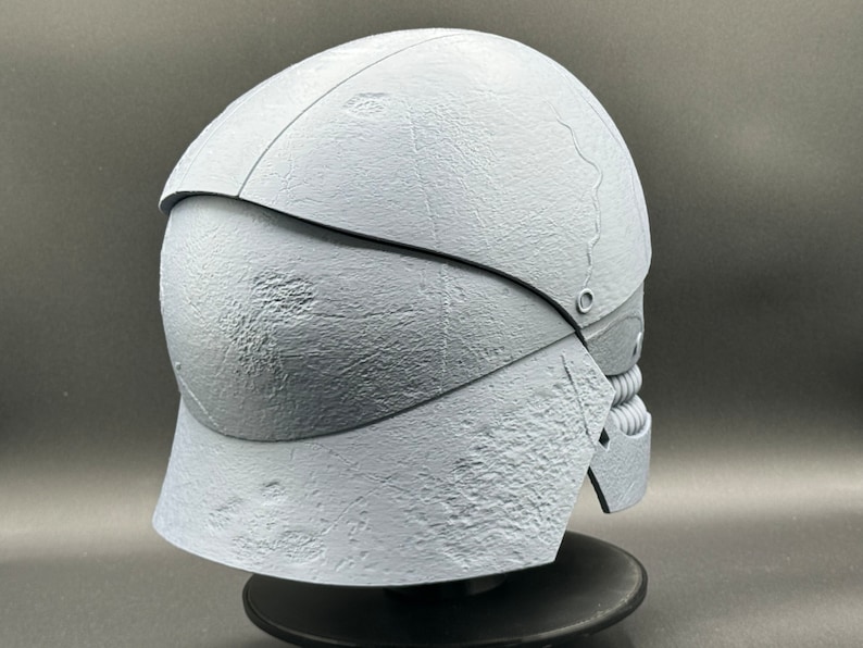 May include: A white, textured helmet with a segmented design. The helmet has a rounded top and a pointed chin guard. The helmet is on a black stand.