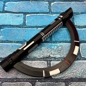 May include: A black and silver lightsaber prop with a curved, segmented accessory. The lightsaber features a detailed handle and a cylindrical design. The accessory is a dark gray with white and red accents. The items are set against a blue brick background.