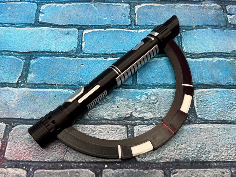 May include: A black and silver lightsaber prop with a curved, segmented accessory. The lightsaber features a detailed handle and a cylindrical design. The accessory is a dark gray with white and red accents. The items are set against a blue brick background.