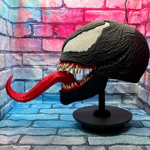 May include: A black Venom mask with a textured surface, white eye details, and a long red tongue. The mask is displayed on a black stand, showcasing the character's menacing features. The mask is a collectible item.