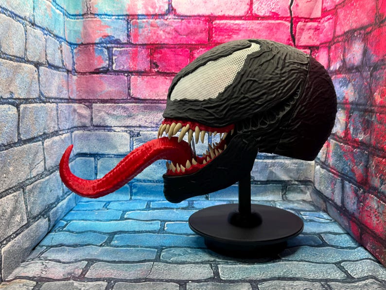May include: A black Venom mask with a textured surface, white eye details, and a long red tongue. The mask is displayed on a black stand, showcasing the character's menacing features. The mask is a collectible item.