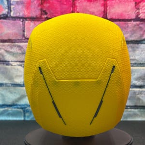 Puede incluir: Casco deportivo amarillo brillante con superficie texturizada y l&iacute;neas negras. El casco se muestra sobre un fondo de pared de ladrillos.