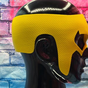 K&ouml;nnte beinhalten: Eine leuchtend gelbe Superheldenmaske mit texturierter Oberfl&auml;che und spitzem Design, das Stirn und Schl&auml;fen bedeckt, getragen auf einem gl&auml;nzend schwarzen Schaufensterpuppenkopf. Die Maske hat ausgepr&auml;gte Augenausschnitte und eine konturierte Passform.