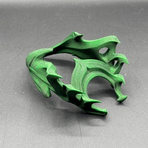 Puede incluir: Una escultura abstracta verde, impresa en 3D, con un dise&ntilde;o org&aacute;nico complejo. El objeto tiene una serie de formas curvas en forma de hoja y bordes puntiagudos. La escultura se muestra sobre una superficie gris.