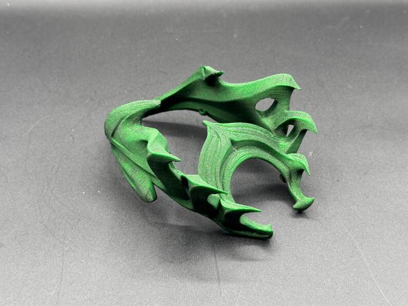 Puede incluir: Una escultura abstracta verde, impresa en 3D, con un dise&ntilde;o org&aacute;nico complejo. El objeto tiene una serie de formas curvas en forma de hoja y bordes puntiagudos. La escultura se muestra sobre una superficie gris.