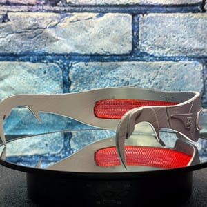 May include: A pair of silver-colored, curved metal accessories with a red, rectangular accent. The accessories are displayed on a reflective surface, with a blue and gray brick wall in the background. The design features a hook-like shape.