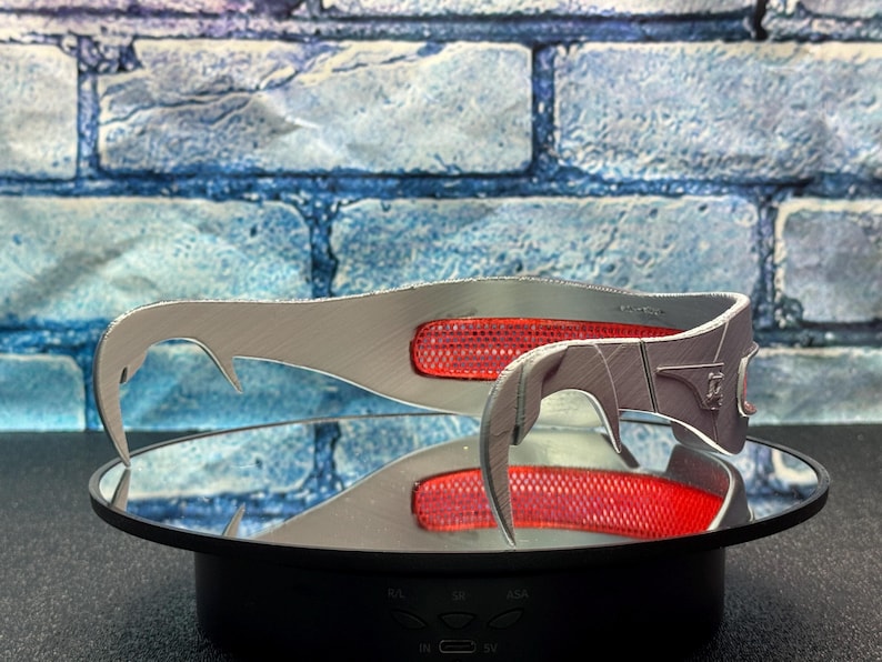 May include: A pair of silver-colored, curved metal accessories with a red, rectangular accent. The accessories are displayed on a reflective surface, with a blue and gray brick wall in the background. The design features a hook-like shape.
