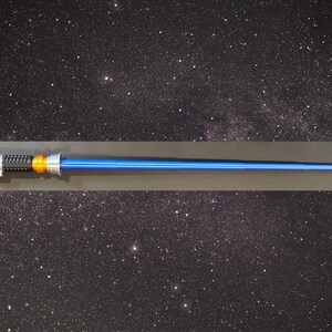 May include: A blue lightsaber prop with silver, black, and orange detailing. The prop features a long, cylindrical blue blade and a detailed handle. The background is a starry night sky.