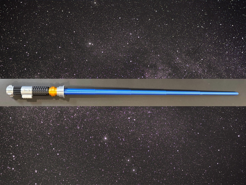 May include: A blue lightsaber prop with silver, black, and orange detailing. The prop features a long, cylindrical blue blade and a detailed handle. The background is a starry night sky.