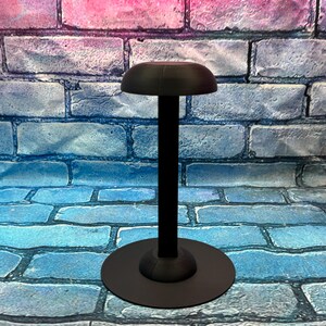 May include: A black hat stand with a round base and a tall, slender pole. The top of the stand has a curved, hat-shaped design. The stand is made of metal and is designed to hold a hat.