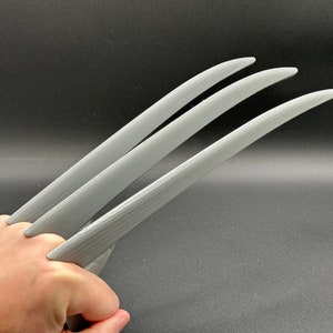 May include: Three silver-colored claws, resembling Wolverine's, are held in a hand. The claws are made of a smooth, slightly textured material.