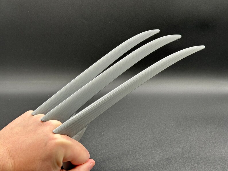 May include: Three silver-colored claws, resembling Wolverine's, are held in a hand. The claws are made of a smooth, slightly textured material.