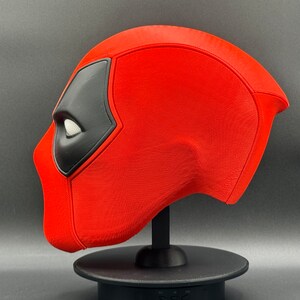 K&ouml;nnte beinhalten: Eine rote und schwarze Deadpool-Maske auf einem schwarzen St&auml;nder. Die Maske besteht aus einem strukturierten Material und hat ein wei&szlig;es Auge.