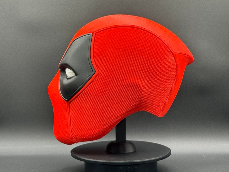 K&ouml;nnte beinhalten: Eine rote und schwarze Deadpool-Maske auf einem schwarzen St&auml;nder. Die Maske besteht aus einem strukturierten Material und hat ein wei&szlig;es Auge.