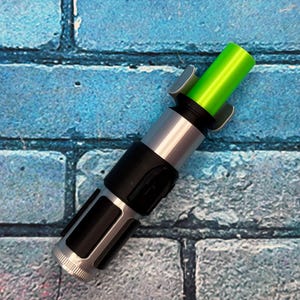 May include: A toy lightsaber with a green blade and a silver and black handle. The hilt has a detailed design with a cylindrical body and a textured grip. The green blade is cylindrical and glows brightly. The toy is set against a blue brick wall.