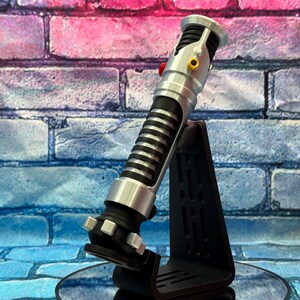 May include: A silver and black lightsaber prop with a ribbed handle and a red button, displayed on a black stand. The prop has silver accents and a black base. The background is a brick wall with blue, pink, and purple hues.