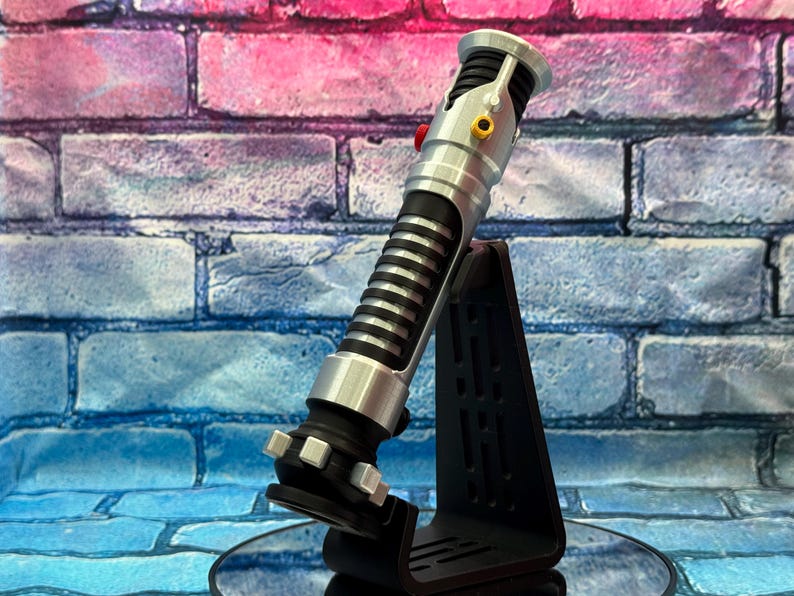 May include: A silver and black lightsaber prop with a ribbed handle and a red button, displayed on a black stand. The prop has silver accents and a black base. The background is a brick wall with blue, pink, and purple hues.