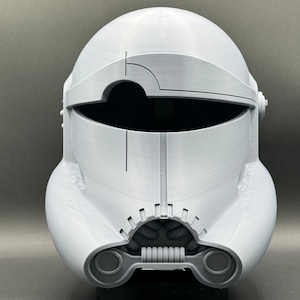 May include: A white plastic clone trooper helmet with a black visor and gray details. The helmet has a textured surface and a design on the front.