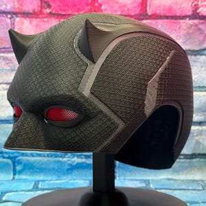Puede incluir: Un casco negro con lentes oculares rojas y dos peque&ntilde;os cuernos. El casco tiene una superficie texturizada y se exhibe sobre un soporte negro. El dise&ntilde;o incluye detalles en gris y una nariz puntiaguda.