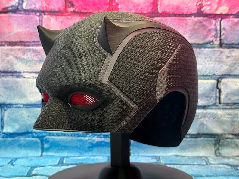 Puede incluir: Un casco negro con lentes oculares rojas y dos peque&ntilde;os cuernos. El casco tiene una superficie texturizada y se exhibe sobre un soporte negro. El dise&ntilde;o incluye detalles en gris y una nariz puntiaguda.