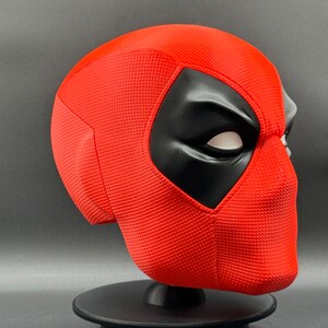 Puede incluir: Una m&aacute;scara de Deadpool roja y negra con una superficie texturizada. La m&aacute;scara est&aacute; en un soporte negro.