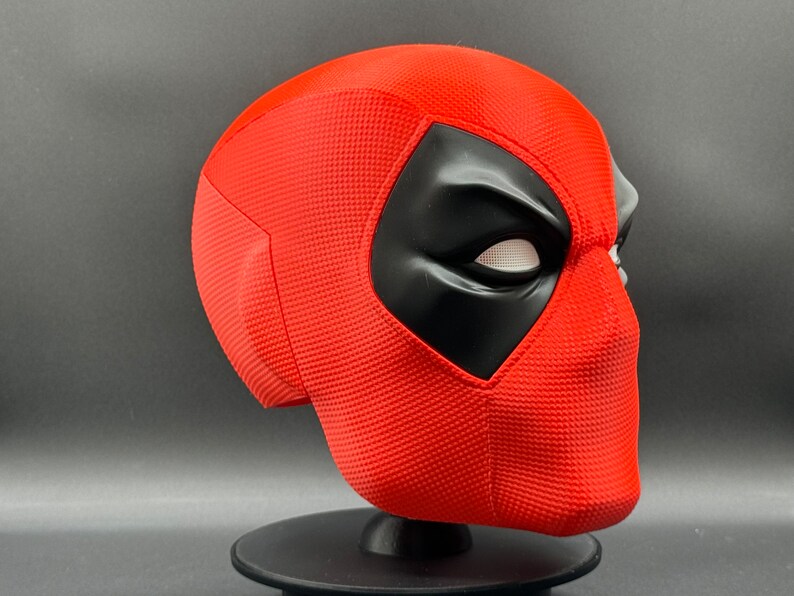 Puede incluir: Una m&aacute;scara de Deadpool roja y negra con una superficie texturizada. La m&aacute;scara est&aacute; en un soporte negro.