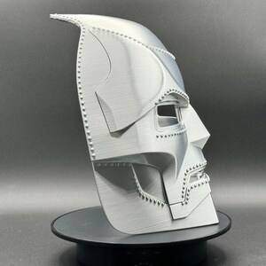 May include: A 3D printed, white, plastic mask with a detailed design. The mask has a pointed top and a textured surface. The mask is designed to resemble a character from a comic book or movie.