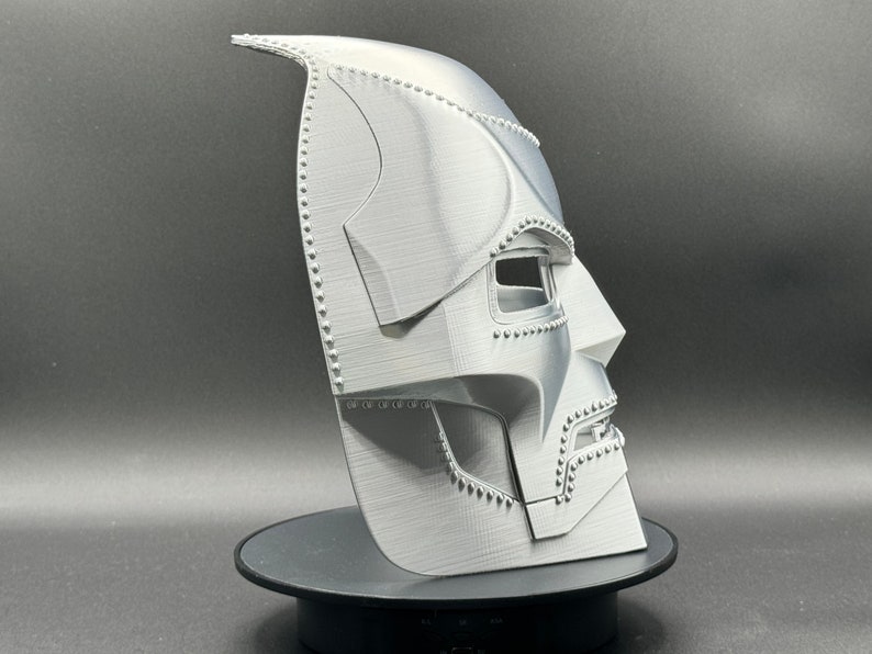 May include: A 3D printed, white, plastic mask with a detailed design. The mask has a pointed top and a textured surface. The mask is designed to resemble a character from a comic book or movie.