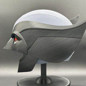 May include: A black and gray 3D printed helmet with a textured surface and red eyes. The helmet is on a black stand.