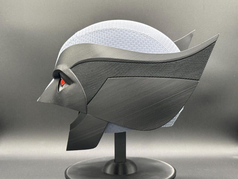 May include: A black and gray 3D printed helmet with a textured surface and red eyes. The helmet is on a black stand.