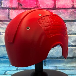 May include: A bright red helmet with a textured design on the top and three silver rivets. The helmet is displayed on a black stand against a brick wall background. The helmet is a cosplay item.