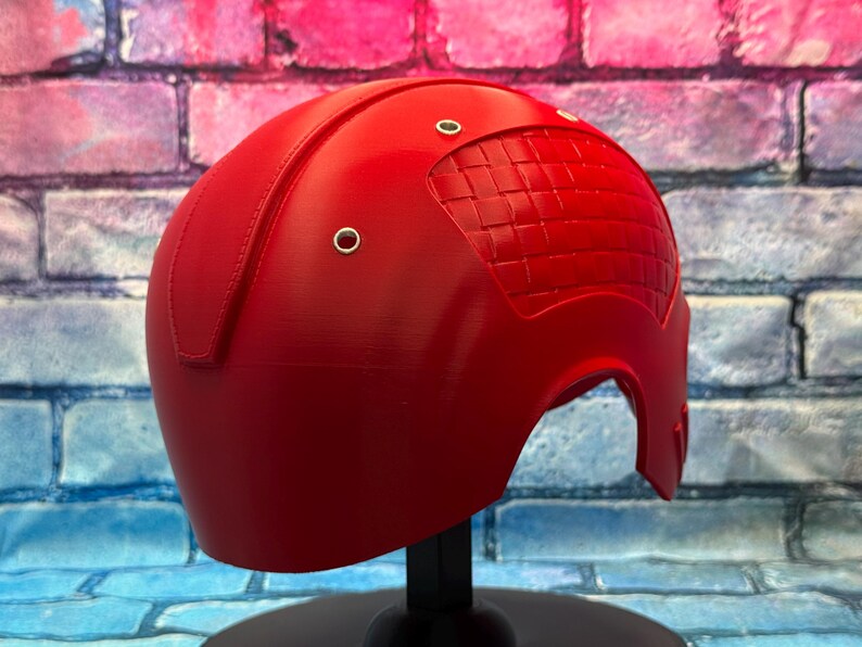 May include: A bright red helmet with a textured design on the top and three silver rivets. The helmet is displayed on a black stand against a brick wall background. The helmet is a cosplay item.