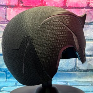 Puede incluir: Un casco negro con una superficie texturizada y un dise&ntilde;o en forma de oreja puntiaguda. El casco se exhibe sobre un soporte negro contra un fondo de pared de ladrillos de colores. El dise&ntilde;o del casco es angular y elegante.