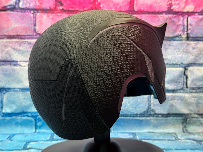 Puede incluir: Un casco negro con una superficie texturizada y un dise&ntilde;o en forma de oreja puntiaguda. El casco se exhibe sobre un soporte negro contra un fondo de pared de ladrillos de colores. El dise&ntilde;o del casco es angular y elegante.
